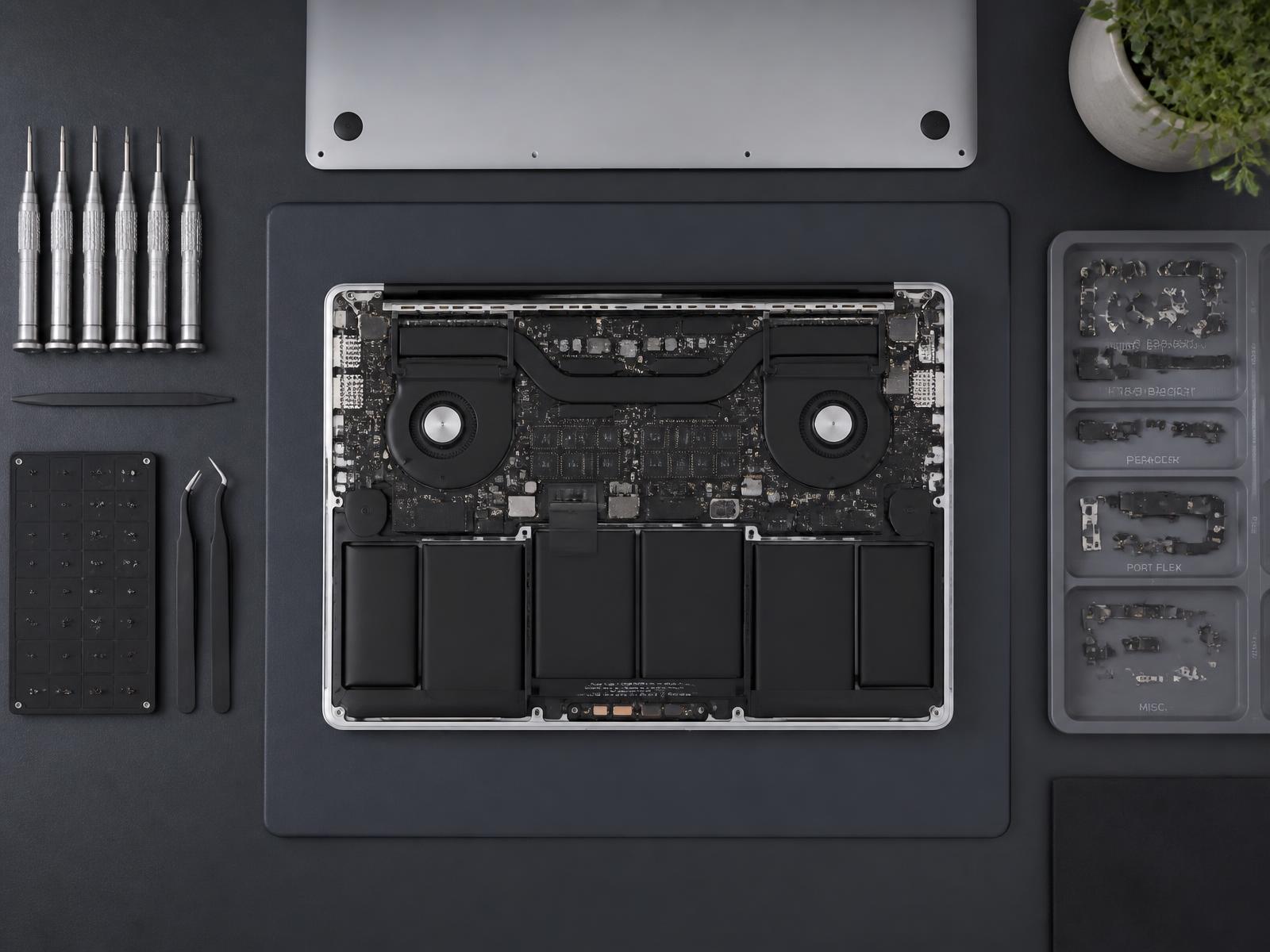 Overhead view of a disassembled MacBook with tools laid out on a graphite workbench